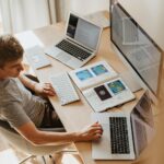 A man multitasks with laptops and a desktop, coding in a home office setting.