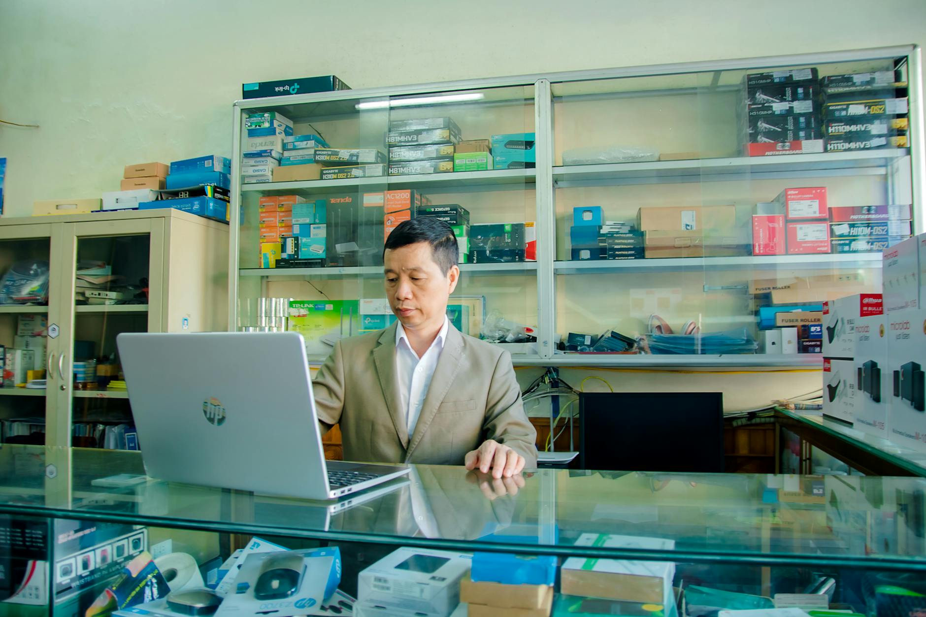 An Asian man using a laptop in an electronics shop with various devices on display.